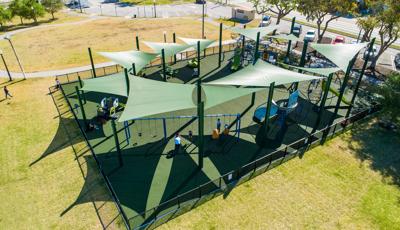 Elevated view of a large fenced in play area with multiple play structures, climbers and swing sets all covered by large green shade sails overhead.