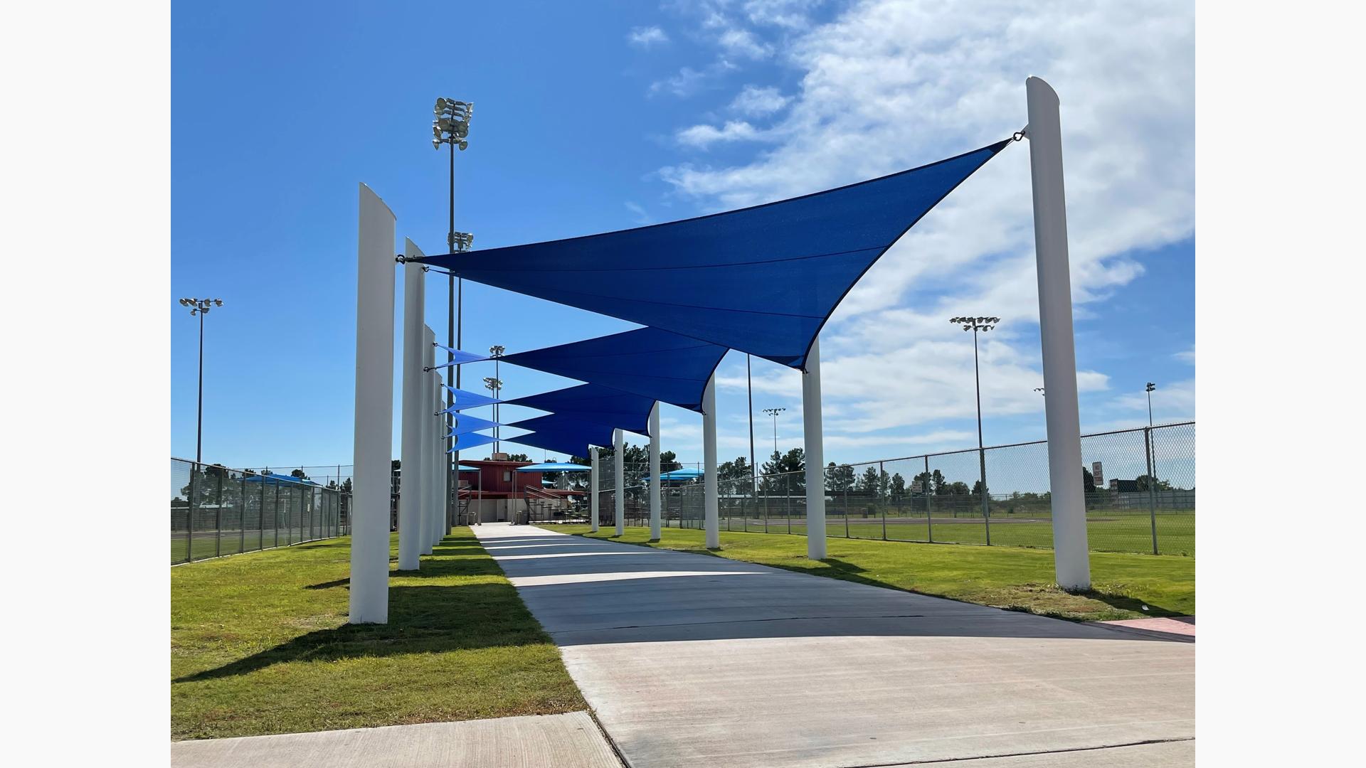 A concrete pathway between two baseball fields has large blue triangular shade sails leading all the way to the complex building in the center of the surrounding ball fields.