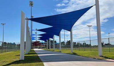 A concrete pathway between two baseball fields has large blue triangular shade sails leading all the way to the complex building in the center of the surrounding ball fields.