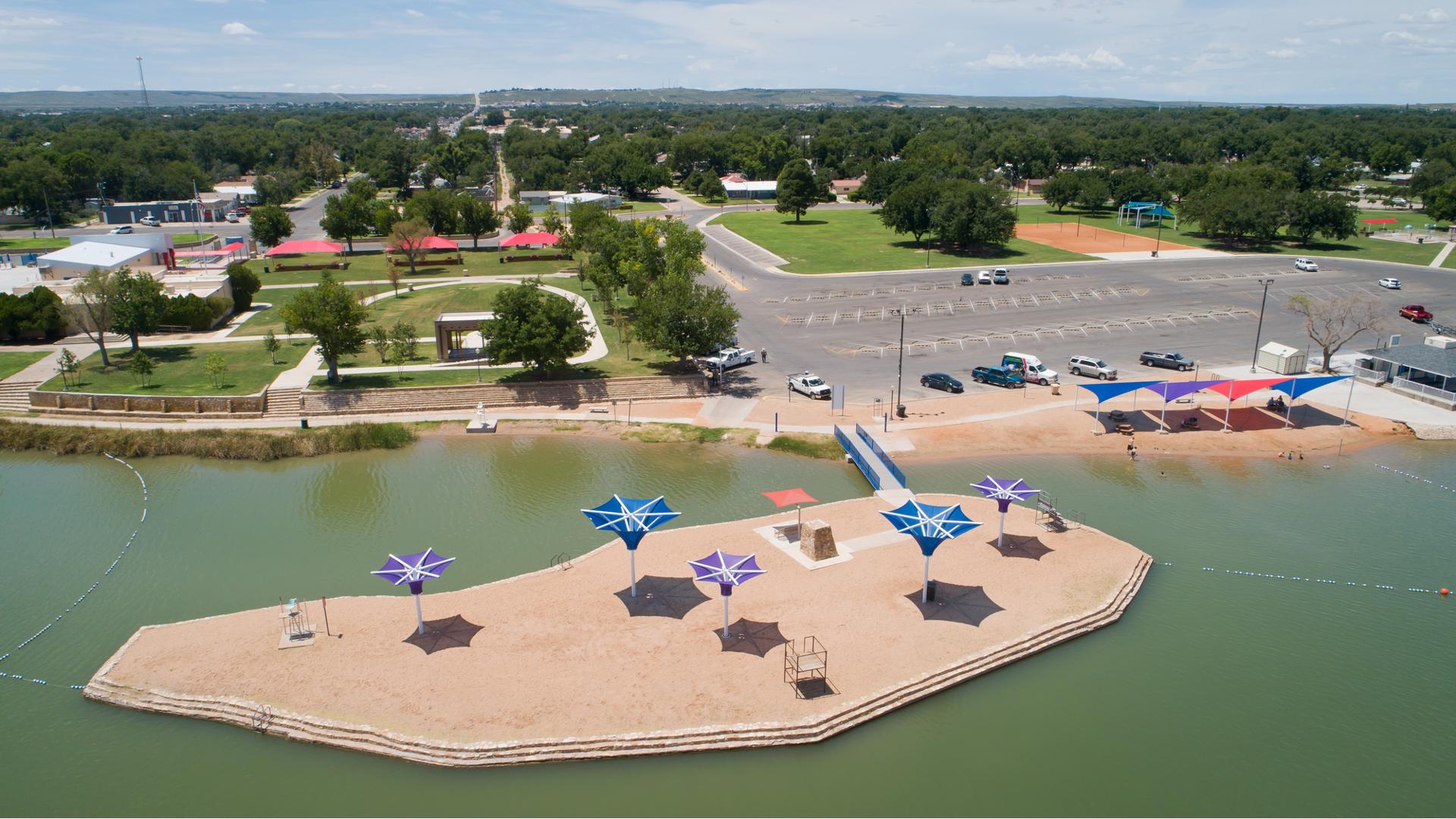 Aerial view of beach area with an island that includes purple and blue shade structures. The beach includes a blue, purple and red colored side-by-side peak shaped shade structures. 