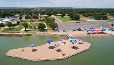 Aerial view of beach area with an island that includes purple and blue shade structures. The beach includes a blue, purple and red colored side-by-side peak shaped shade structures. 