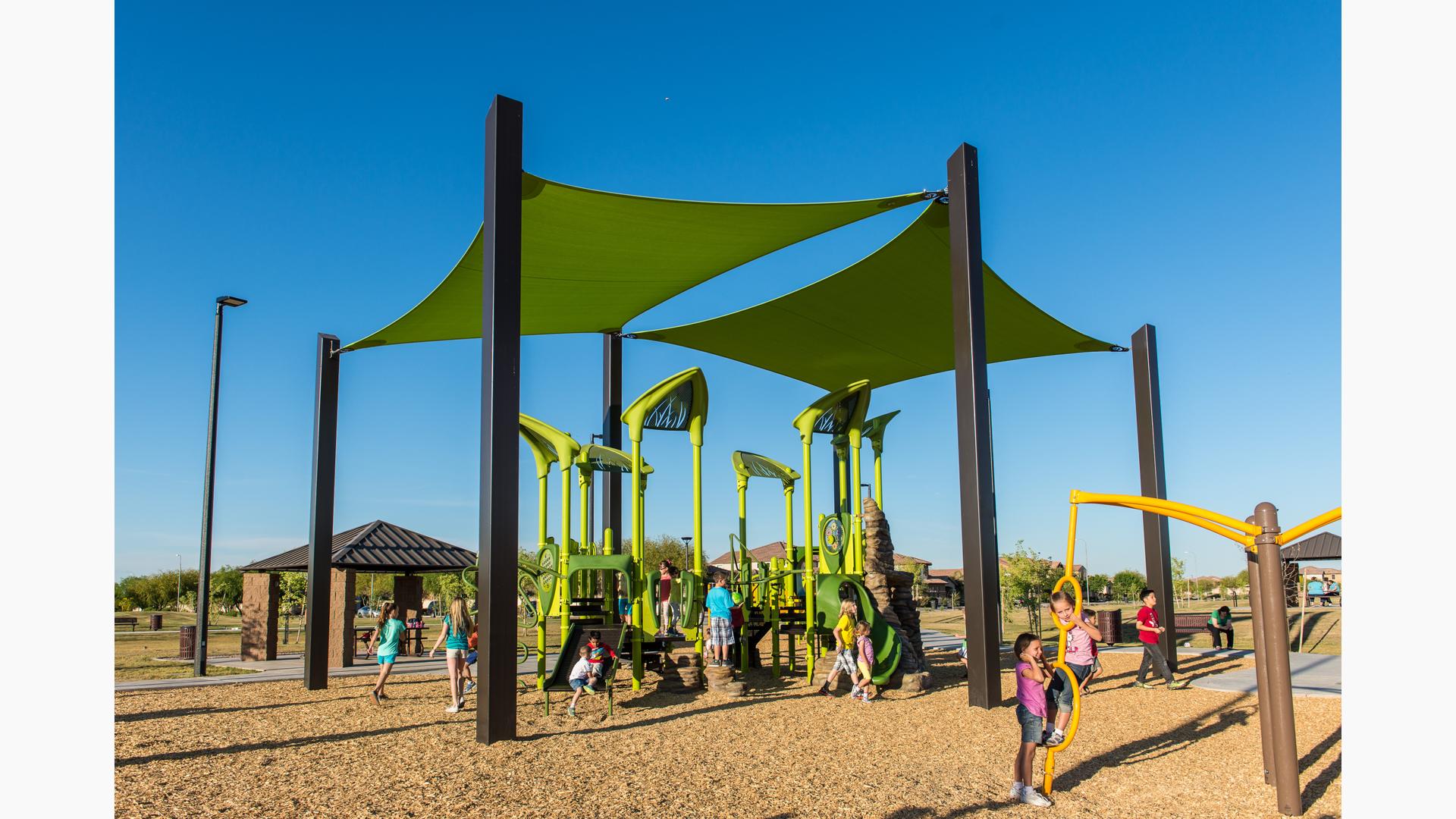 Two girls playing on a Spinner as other children enjoy the natural-themed playground colored bright green with two green shade structures overhead. 