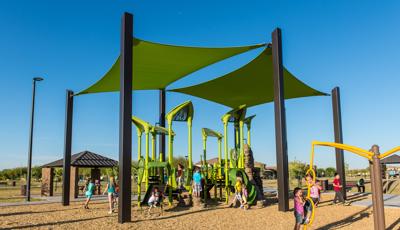 Two girls playing on a Spinner as other children enjoy the natural-themed playground colored bright green with two green shade structures overhead. 