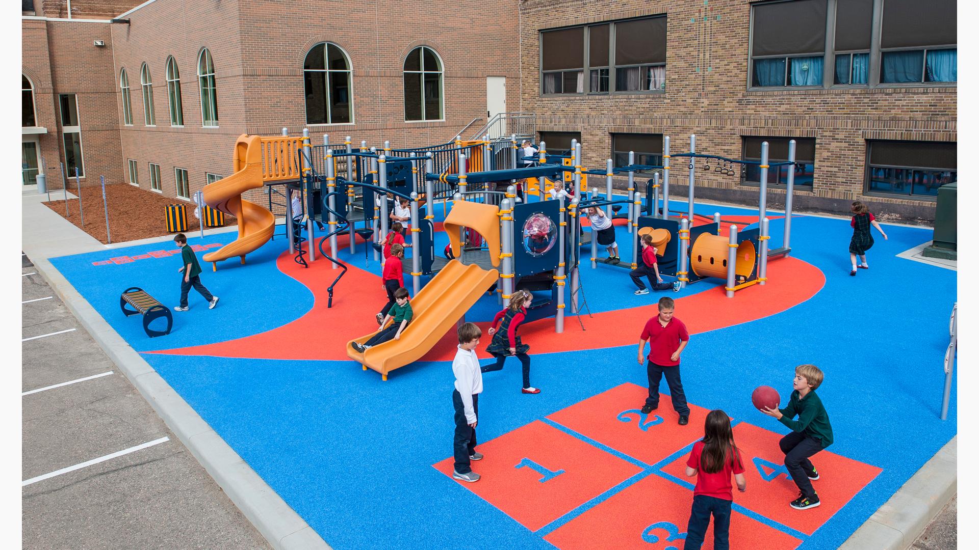 Children playing four-square, while others play on the PlayBooster play system. Saint Maximilian Kolbe Catholic School is in the background.