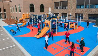 Children playing four-square, while others play on the PlayBooster play system. Saint Maximilian Kolbe Catholic School is in the background.
