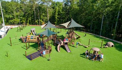 Elevated view of a large play area with green artificial grass contains one large play structure connected with many inclusive accessible ramps, play panels, climbers, and slides. Children play on a manual carousel. The play area is surrounded by a thick wooded area just beyond the playground fencing.