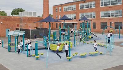 School children in uniforms play on playground against red brick elementary school building in background.