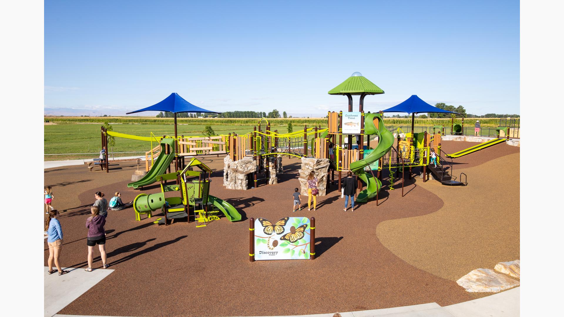 Children and families playing on nature-inspired playground with green slides, bridges, climbing rocks and with interactive panels about the animal life in Idaho. 