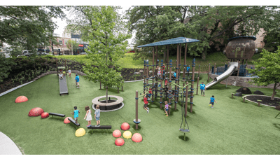 Lots of children playing on a tower net play structure, this one of a kind playground tucked away amongst the trees.  In the foreground a bird nest looking play structure with a stainless steel slide. Kids riding down the Roller slide which is built into the hillside.