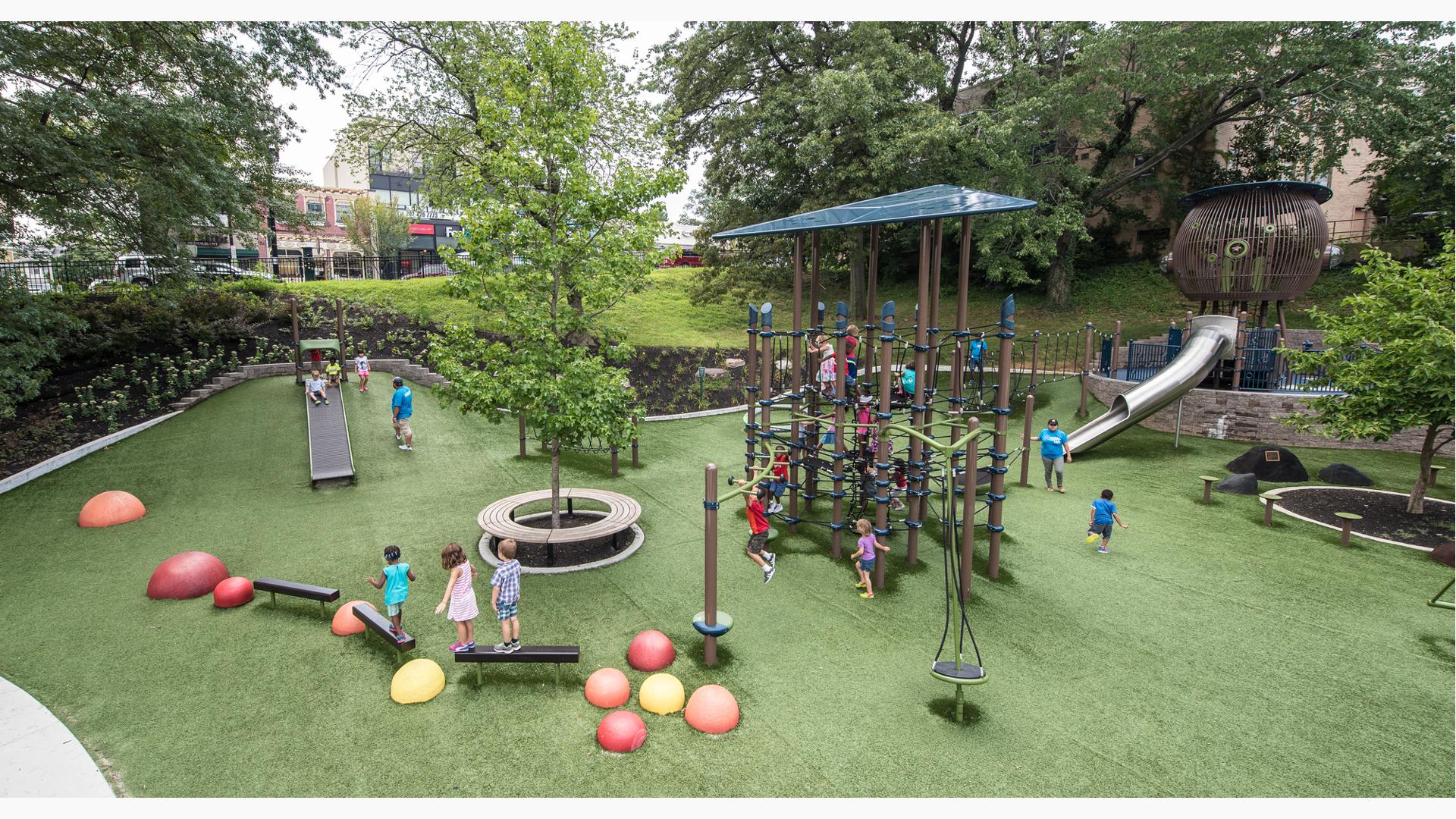 Lots of children playing on a tower net play structure, this one of a kind playground tucked away amongst the trees.  In the foreground a bird nest looking play structure with a stainless steel slide. Kids riding down the Roller slide which is built into the hillside.