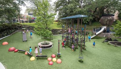 Lots of children playing on a tower net play structure, this one of a kind playground tucked away amongst the trees.  In the foreground a bird nest looking play structure with a stainless steel slide. Kids riding down the Roller slide which is built into the hillside.