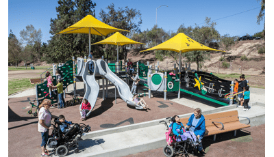 Children enjoying the custom inclusve Ritchie Valens Park. A boy in his wheel chair is helped up the ramp, while another girl sits next to a woman who is assisting her. In the background other children of all abilities ride the down the double slide and two girls are assisted up a climber.