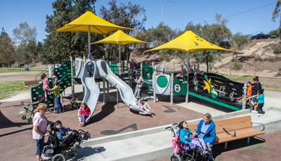 Children enjoying the custom inclusve Ritchie Valens Park. A boy in his wheel chair is helped up the ramp, while another girl sits next to a woman who is assisting her. In the background other children of all abilities ride the down the double slide and two girls are assisted up a climber.