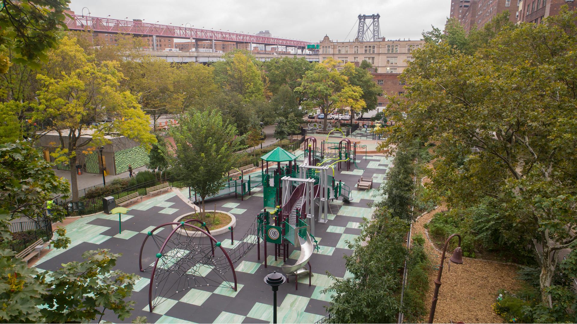 Elevated view of a park amongst city buildings. The playground has a bridge designed like a near by bridge in the distance. 