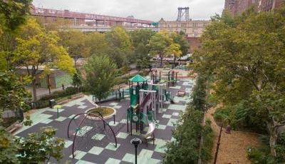 Elevated view of a park amongst city buildings. The playground has a bridge designed like a near by bridge in the distance. 