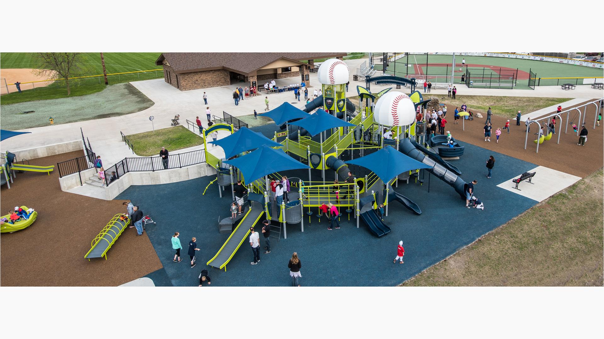Lots of families playing on an inclusive, fully-ramped and accessible playground with four blue shade structures and two baseball roofs.
